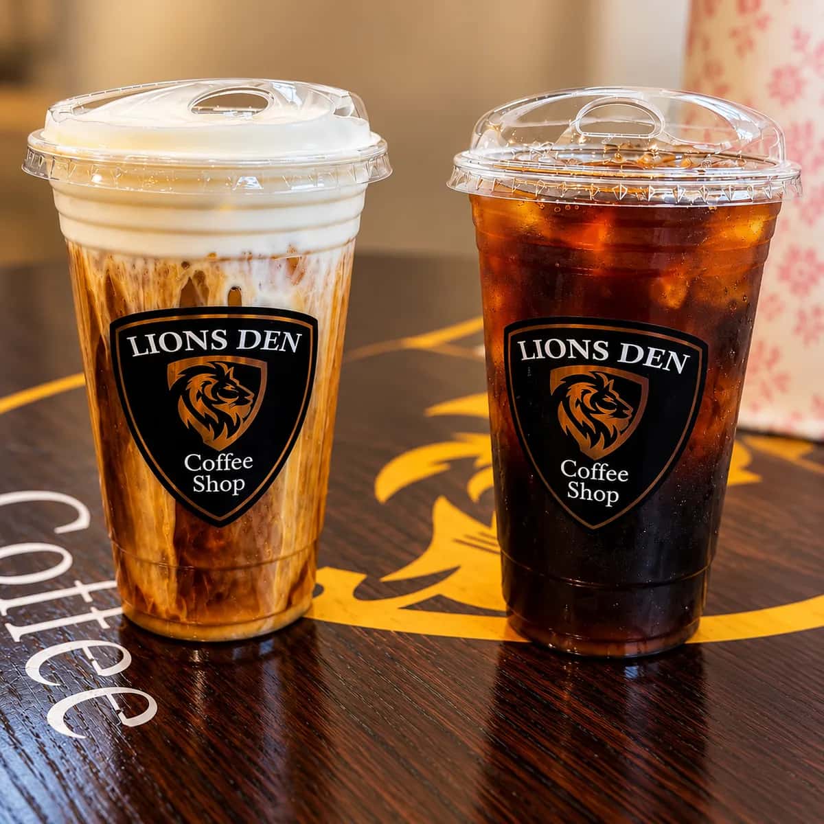 Two iced coffees in Lions Den Coffee Shop cups, one with cream swirled through, one black