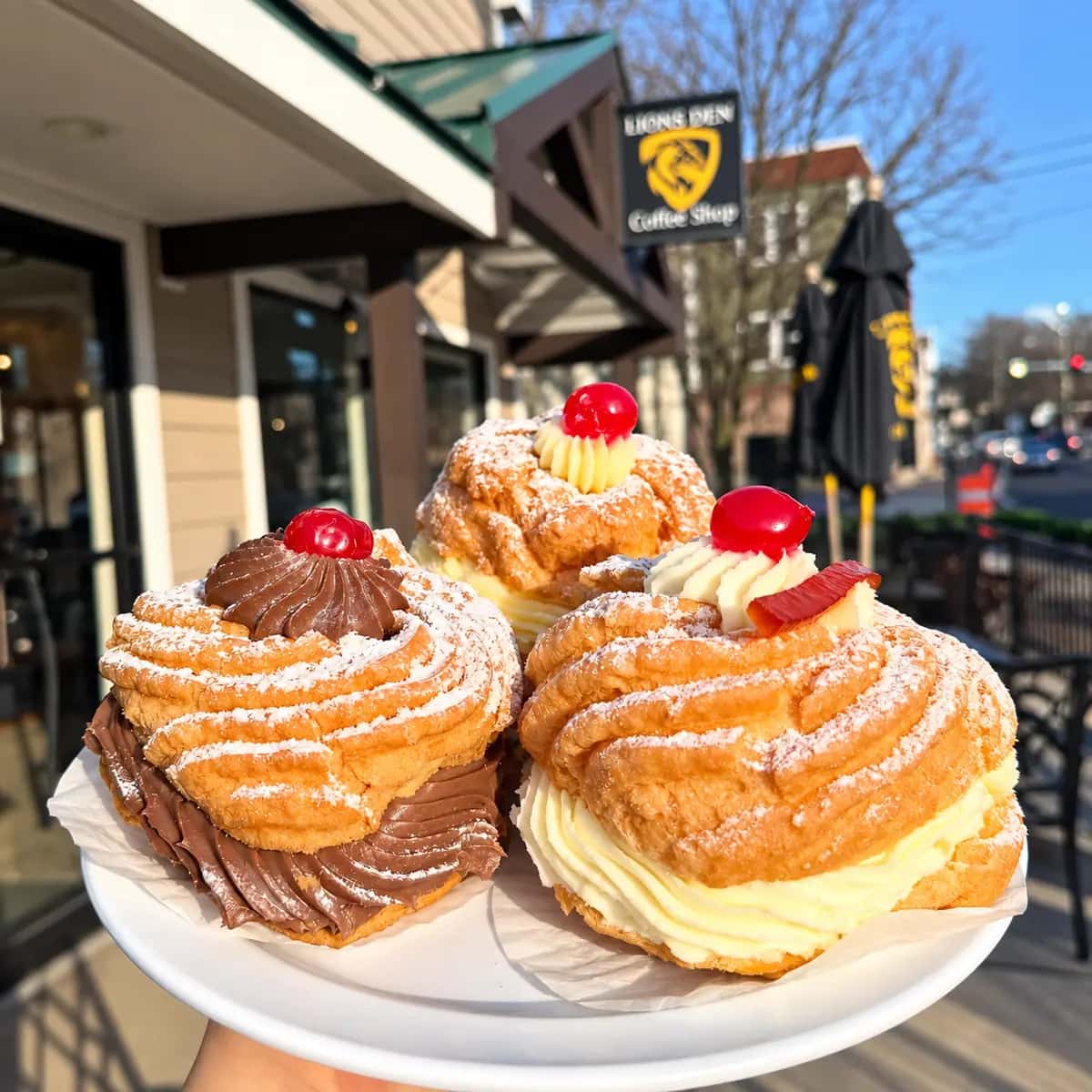 Three Italian cream puffs filled with vanilla and chocolate cream, topped with cherries and powdered sugar, held up outside the Lions Den storefront